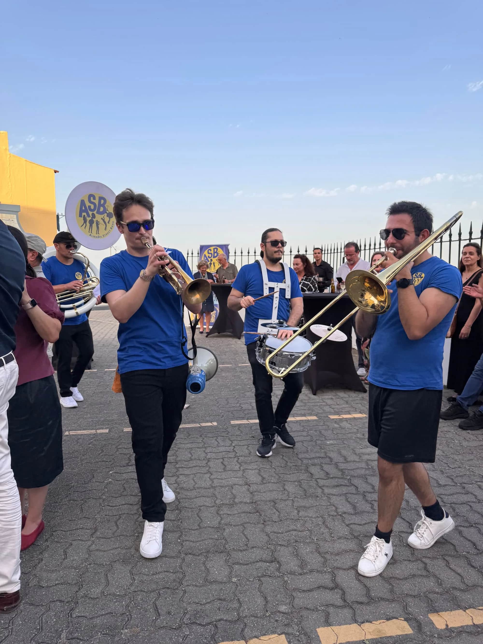 A banda a desfilar por uma calçada, com trompete e trombone, e o público em volta