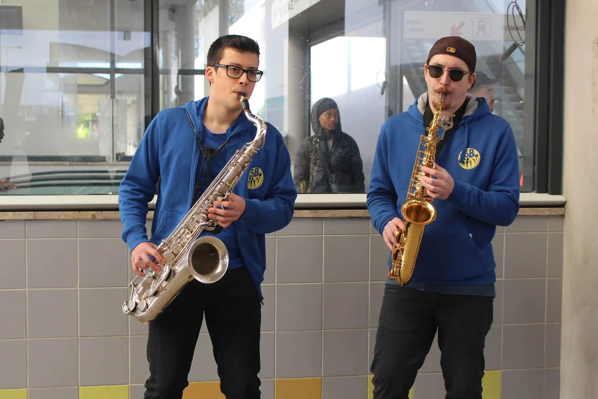 Two saxophone players performing in a station as someone watches from a window