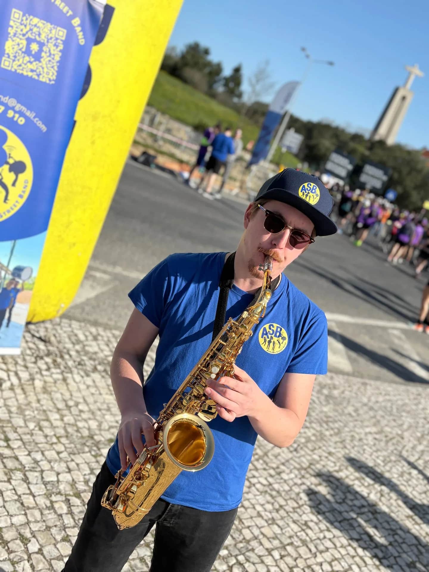 Saxofonista de boné preto e óculos escuros junto à bandeira da ASB
